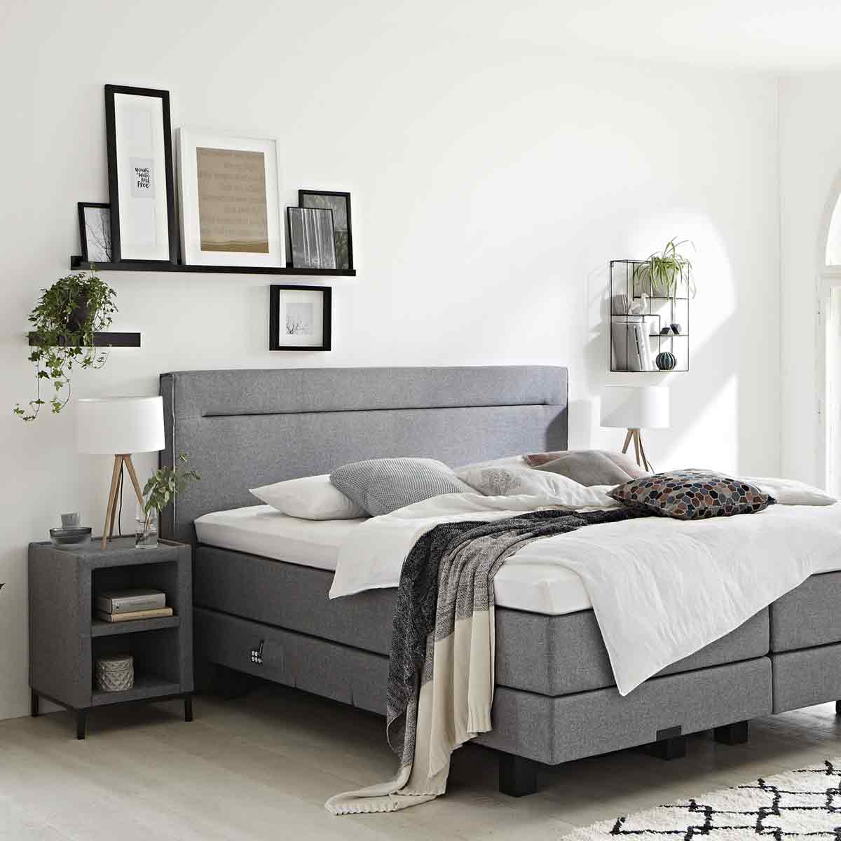 Grijze boxspring met gestoffeerd hoofdbord in een lichte slaapkamer, aangekleed met wit beddengoed en een zachte plaid. Een vrouw ligt ontspannen op bed met een tablet in de hand.