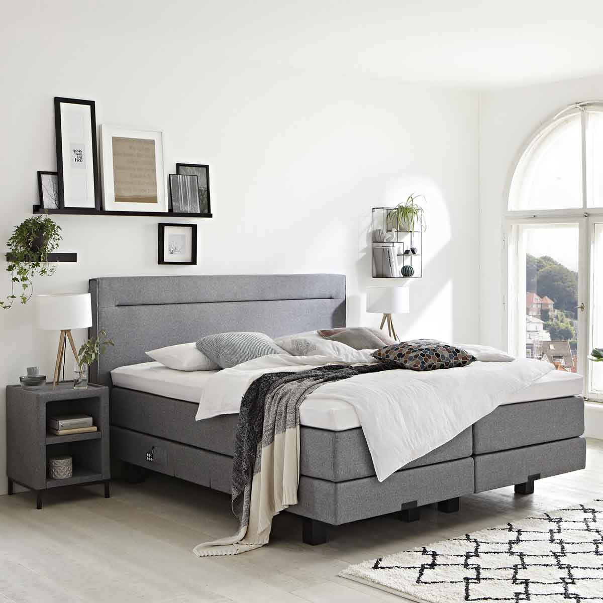 Een grijze boxspring met witte lakens met een zwart wit kleed in een slaapkamer