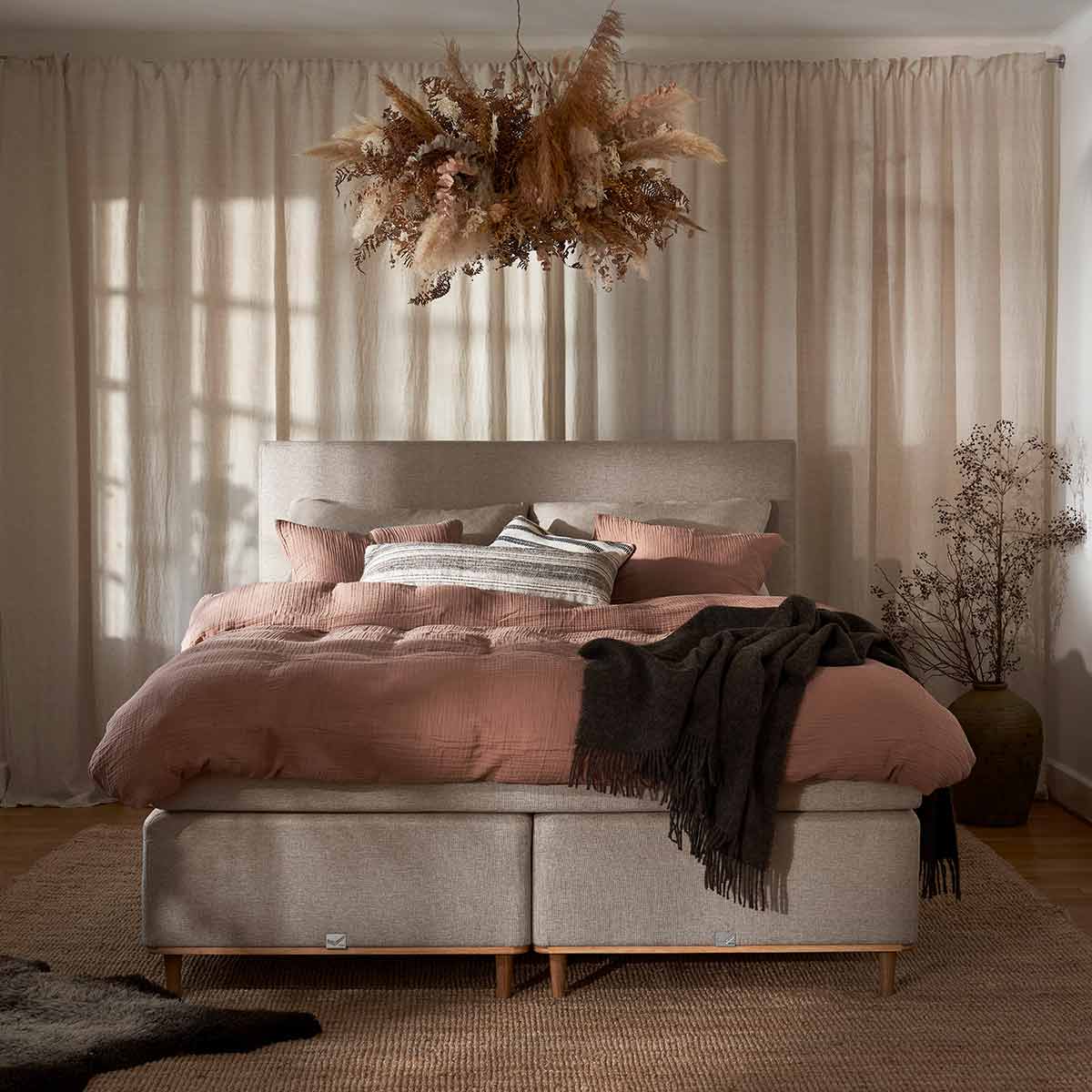 Een romantische slaapkamer met roze beddengoed, linnen boxspring, zachte gordijnen en hanglamp van droogbloemen