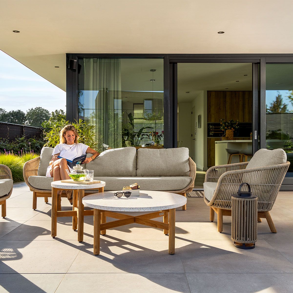 Moderne loungeset op terras met houten details, vrouw lezend en lichte kussens in zonlicht