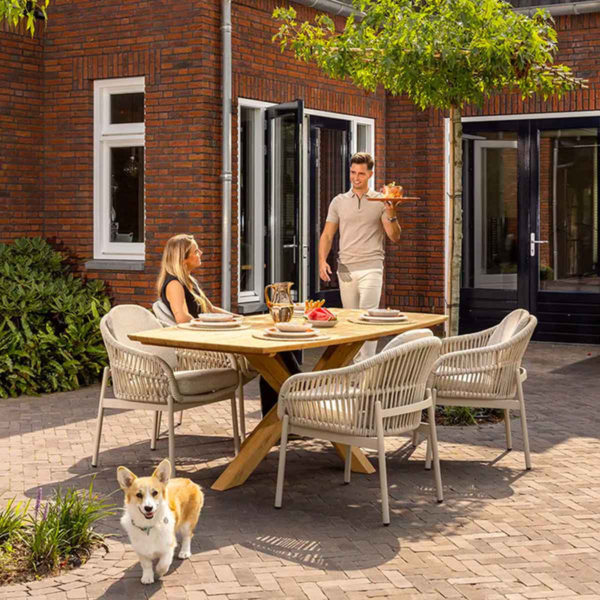 Houten tuintafel met gevlochten stoelen, stel dineert buiten en hond loopt over terras