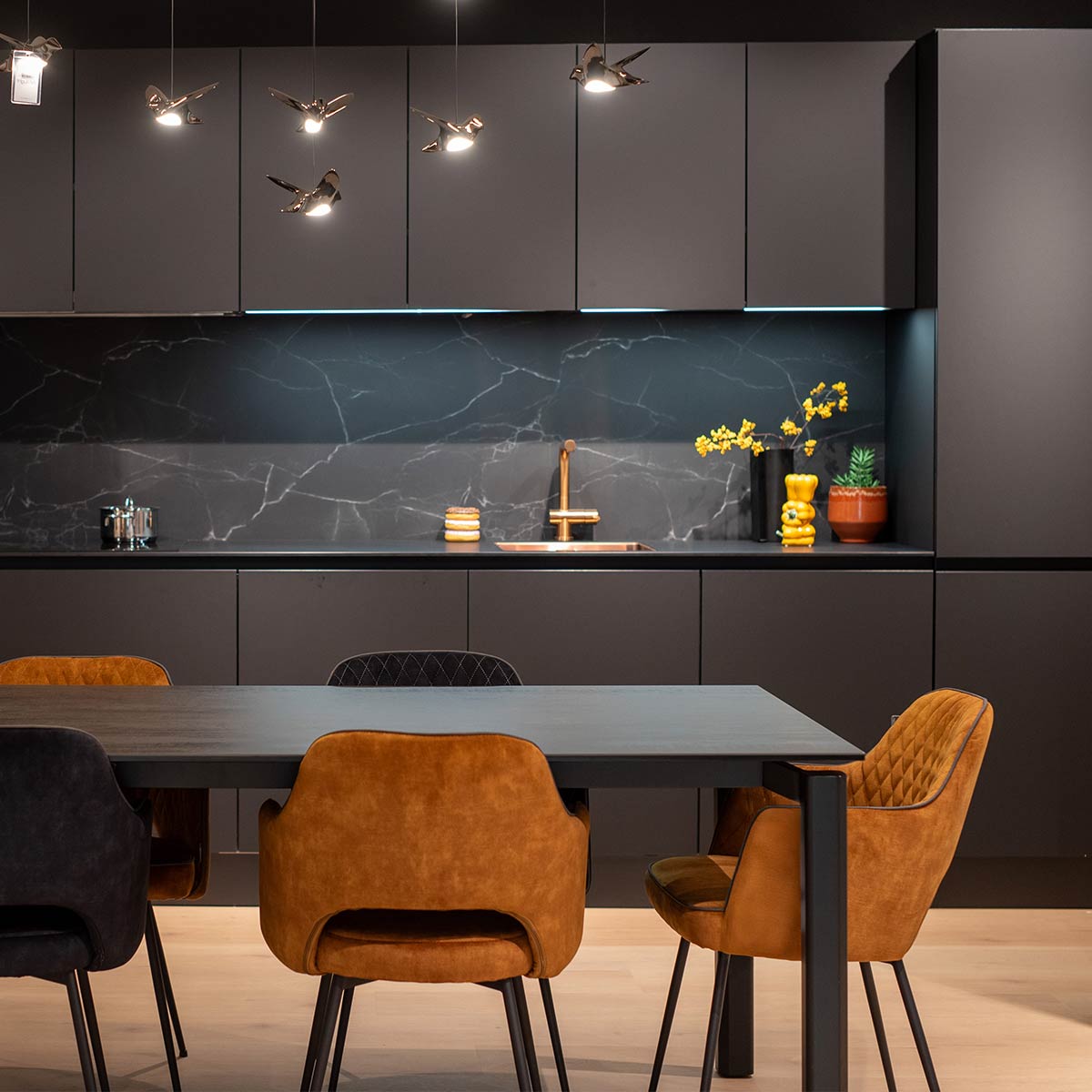 Strakke zwarte keuken met marmeren achterwand, hanglampen, houten tafel en fluwelen eetkamerstoelen