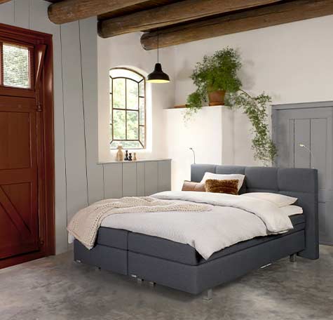 Donkergrijze boxspring van Avek in landelijke slaapkamer met houten balkenplafond, wit beddengoed en plant op de vensterbank