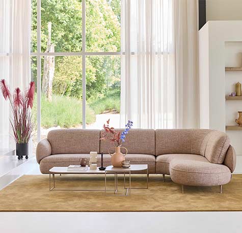 Beige hoekbank van Bert Plantagie met afgeronde vormen in lichte woonkamer met hoge ramen, zacht vloerkleed en moderne salontafel.