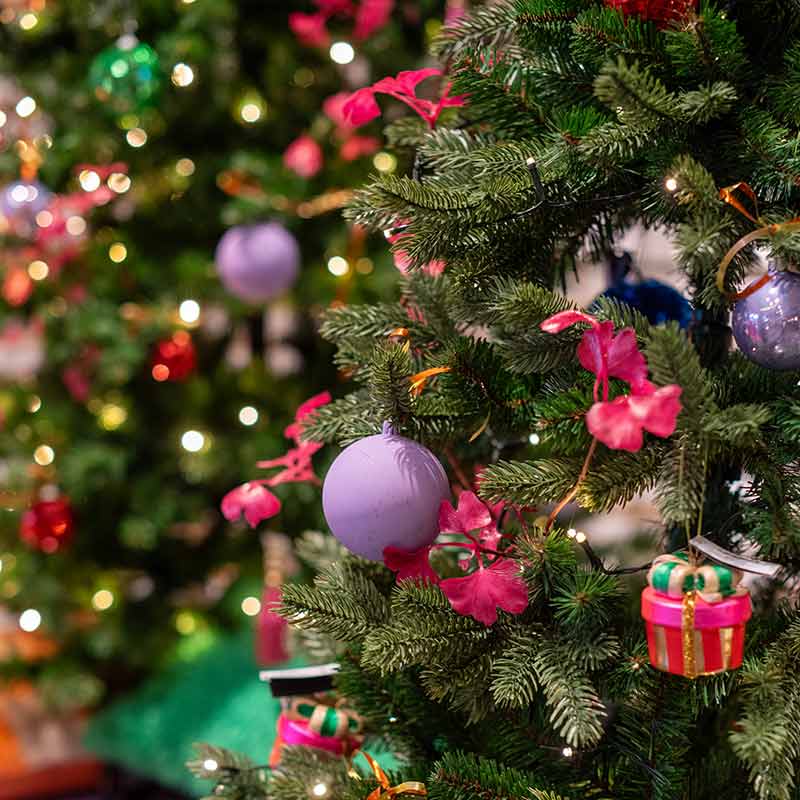 Kleurrijke kerstboom versierd met paarse kerstballen, roze bloemen en speelse ornamenten zoals cadeautjes, met sfeervolle verlichting op de achtergrond