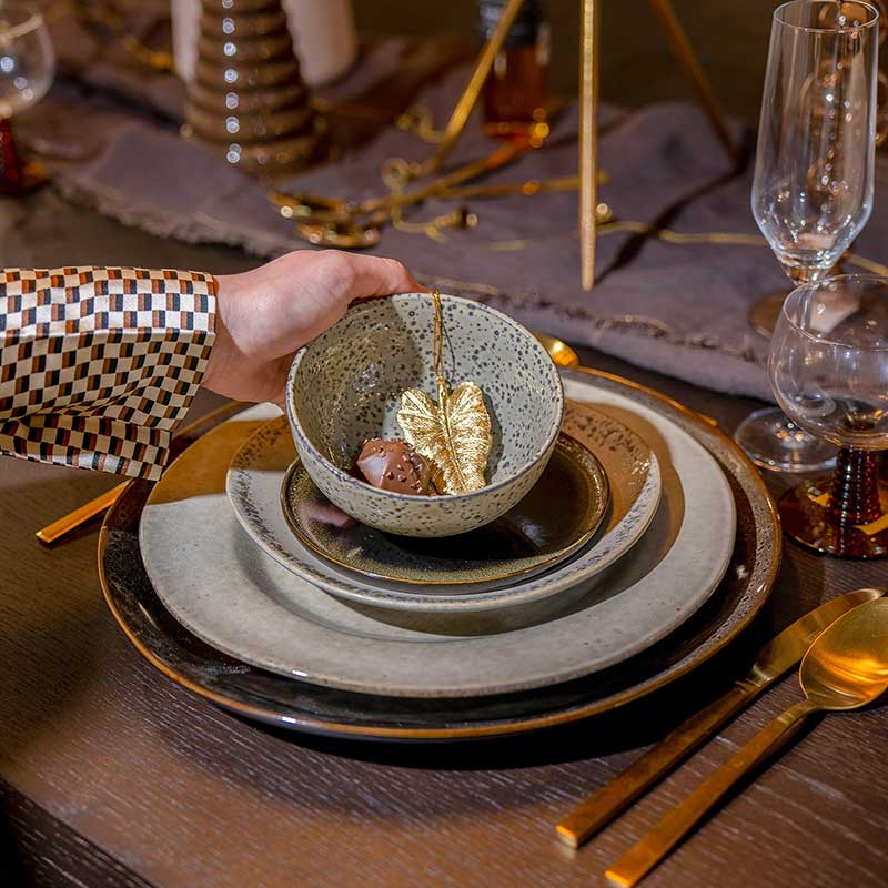Close-up van een hand die een keramische kom met een gouden decoratief blad op een luxe gedekte tafel plaatst, voorzien van aardetinten servies en gouden bestek