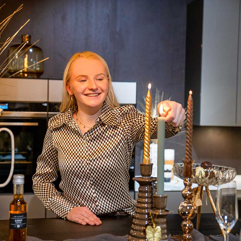 Interieurstyliste van Home Center steekt een decoratieve kaars aan op een feestelijk gedekte kersttafel in een moderne keukenomgeving