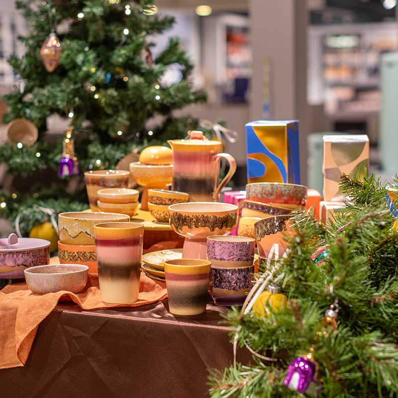 Tafel gedecoreerd met kleurrijk keramisch servies van HKLiving in warme tinten, geplaatst naast een versierde kerstboom in een feestelijke winkelomgeving van Home Center