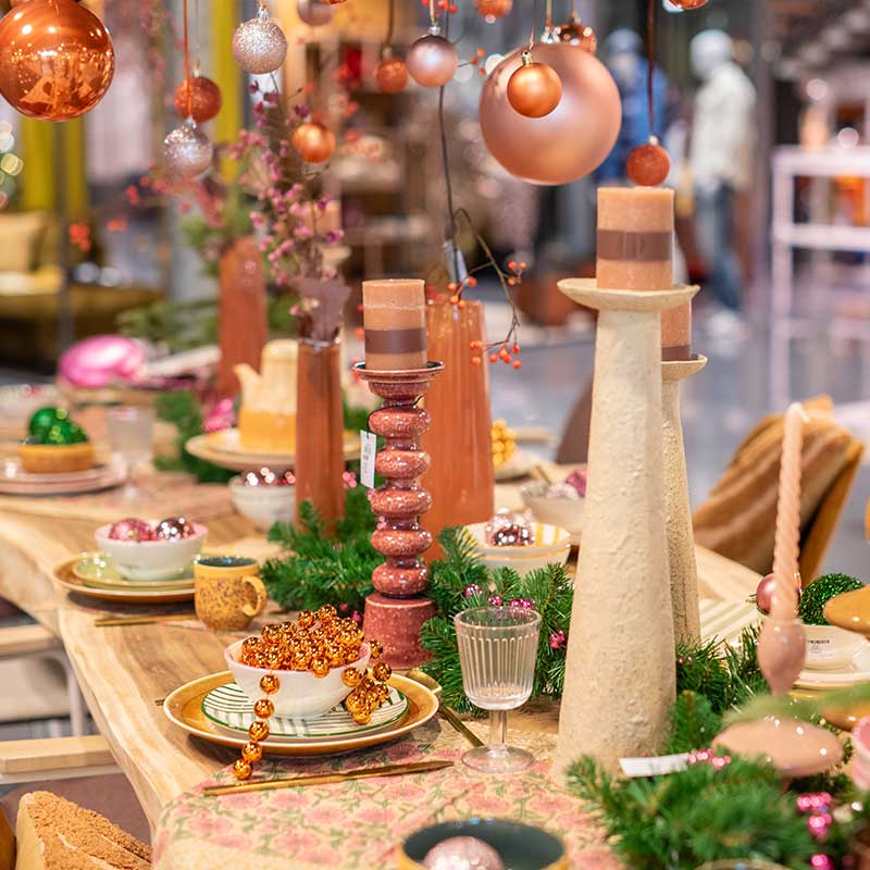 Feestelijk gedekte kersttafel met warme aardetinten, decoratieve kaarsen, kerstballen in oranje en roze tinten, en groene accenten